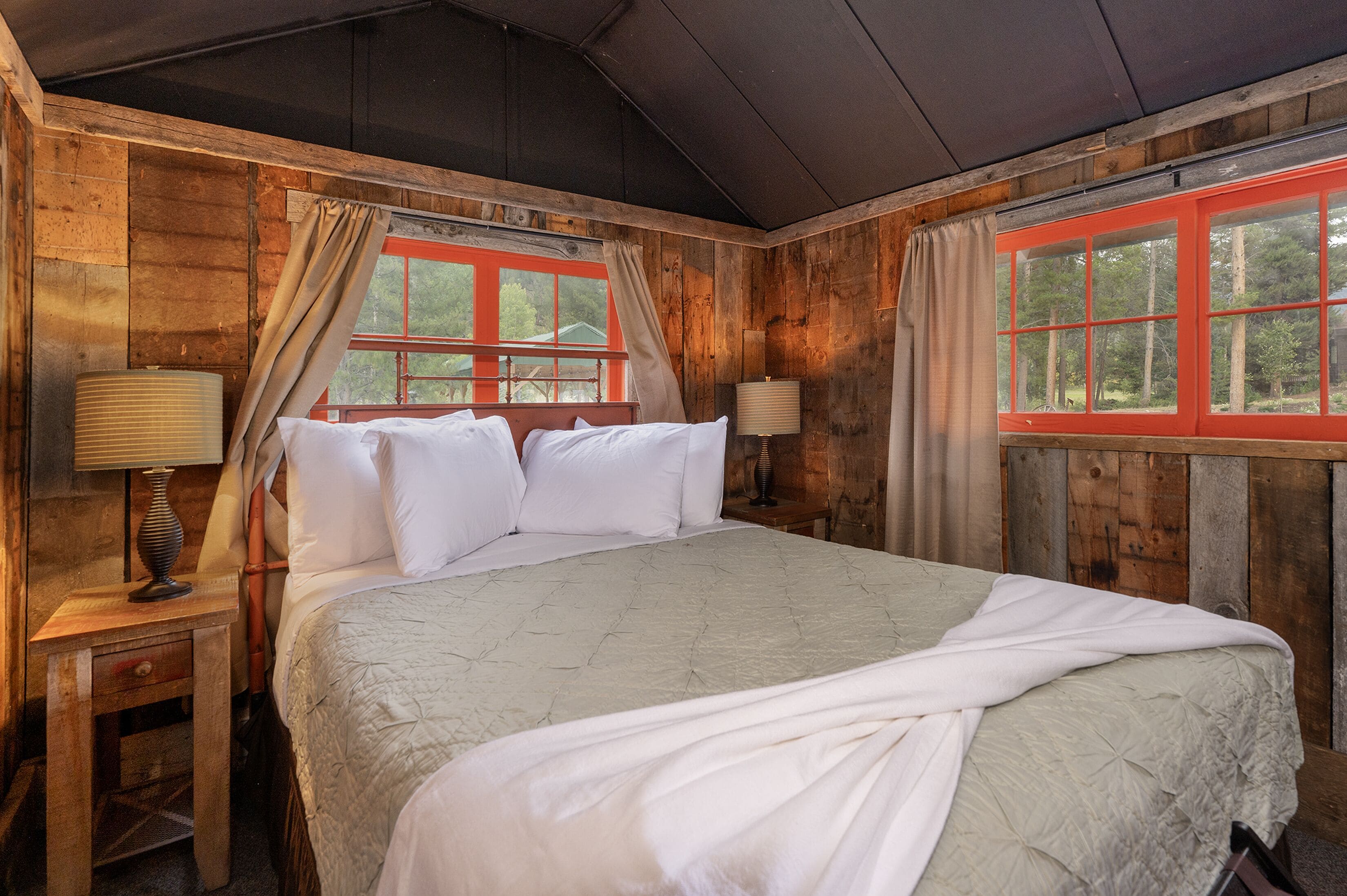 A wooden cabin bedroom with a large bed, two bedside lamps, and a window with red frames.