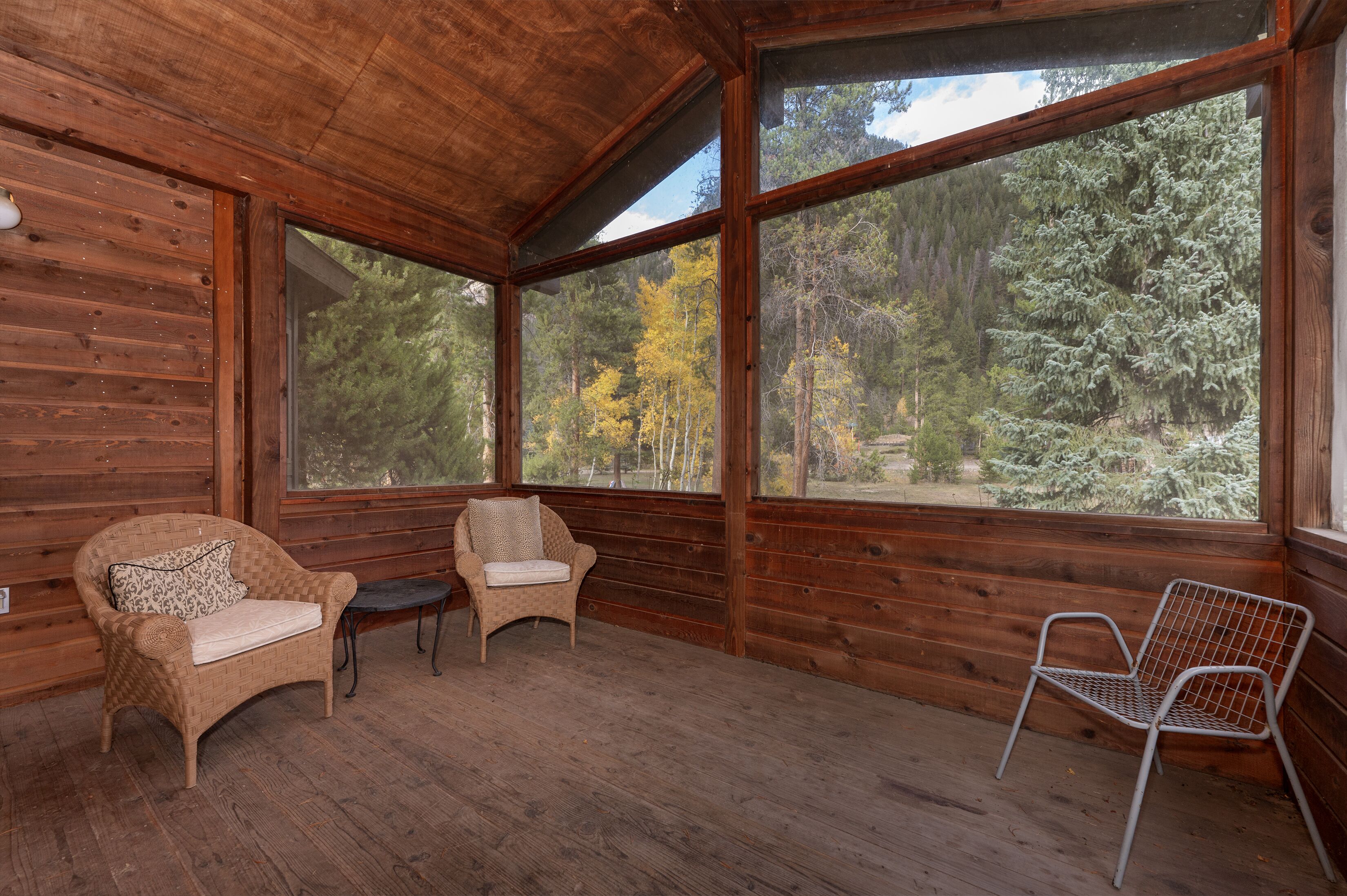 A wooden cabin with large windows, two wicker chairs, and a metal chair.