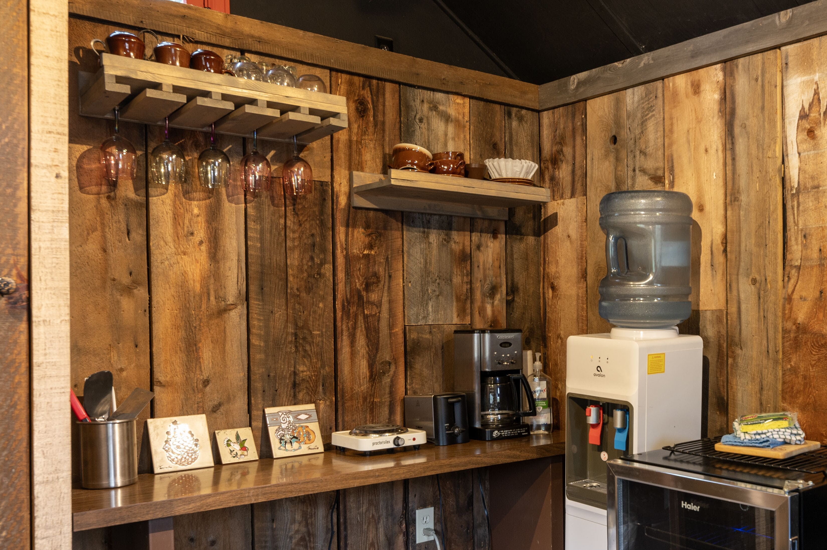 Ett kök med träpanel på väggarna, en kaffemaskin, en vattenautomat och en hylla med muggar och glas.
