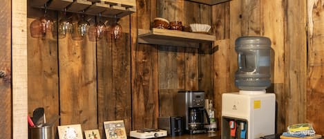 Ett kök med träpanel på väggarna, en kaffemaskin, en vattenautomat och en hylla med muggar och glas.