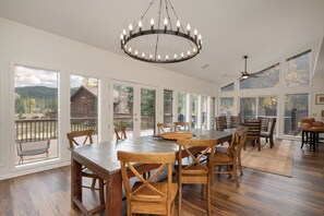 A spacious dining area with a large wooden table and chairs, a chandelier, and large windows offering views of a wooden deck and mountains.
