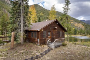 A wooden cabin with a porch and steps leading to the entrance, surrounded by trees and a lake.