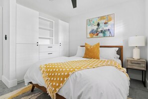Um quarto com cama, mesa de cabeceira, abajur, quadro e guarda-roupa.