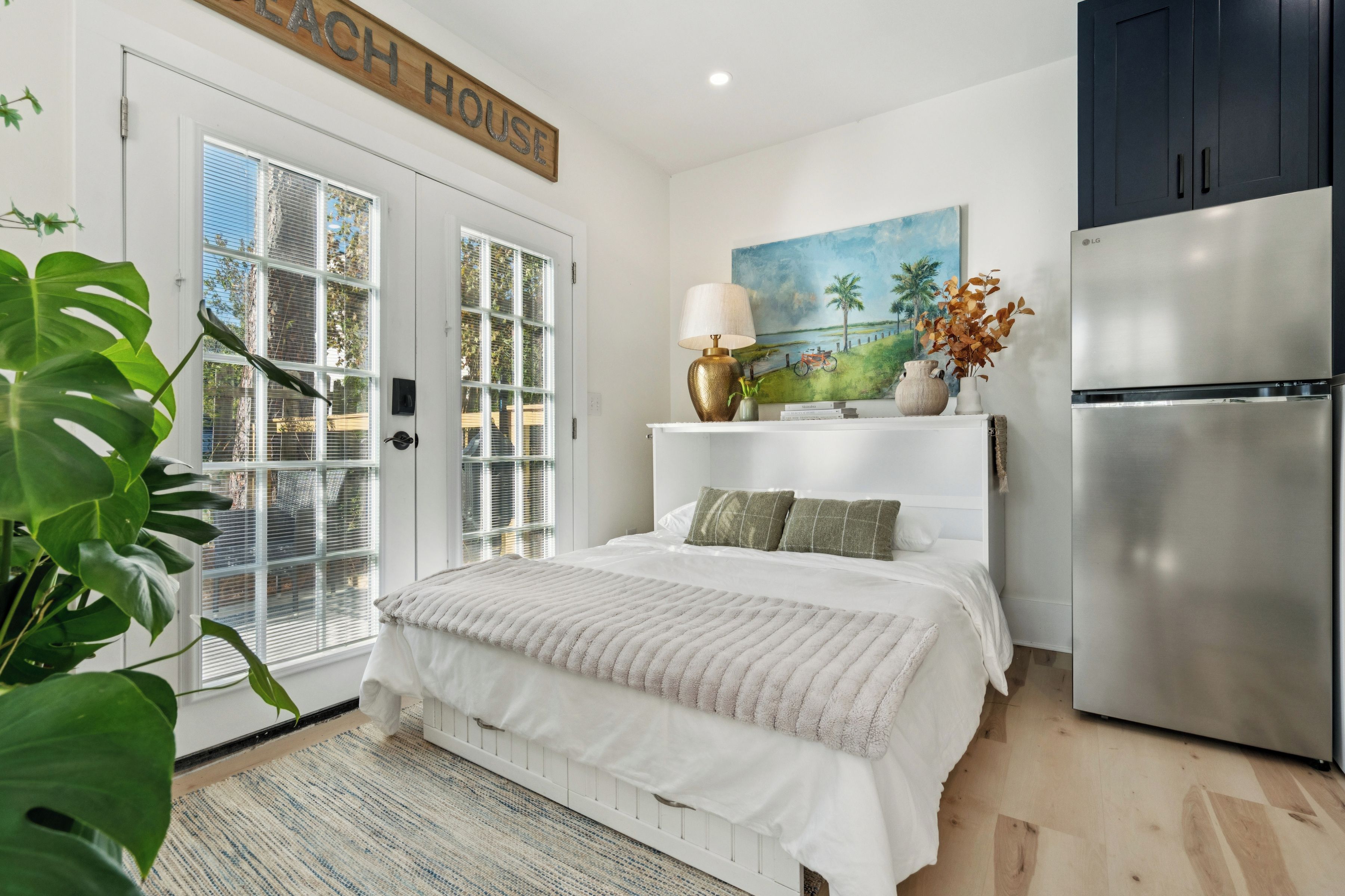 A bedroom with a bed, a refrigerator, a painting, and a plant.