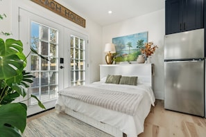 A bedroom with a bed, a refrigerator, a painting, and a plant.