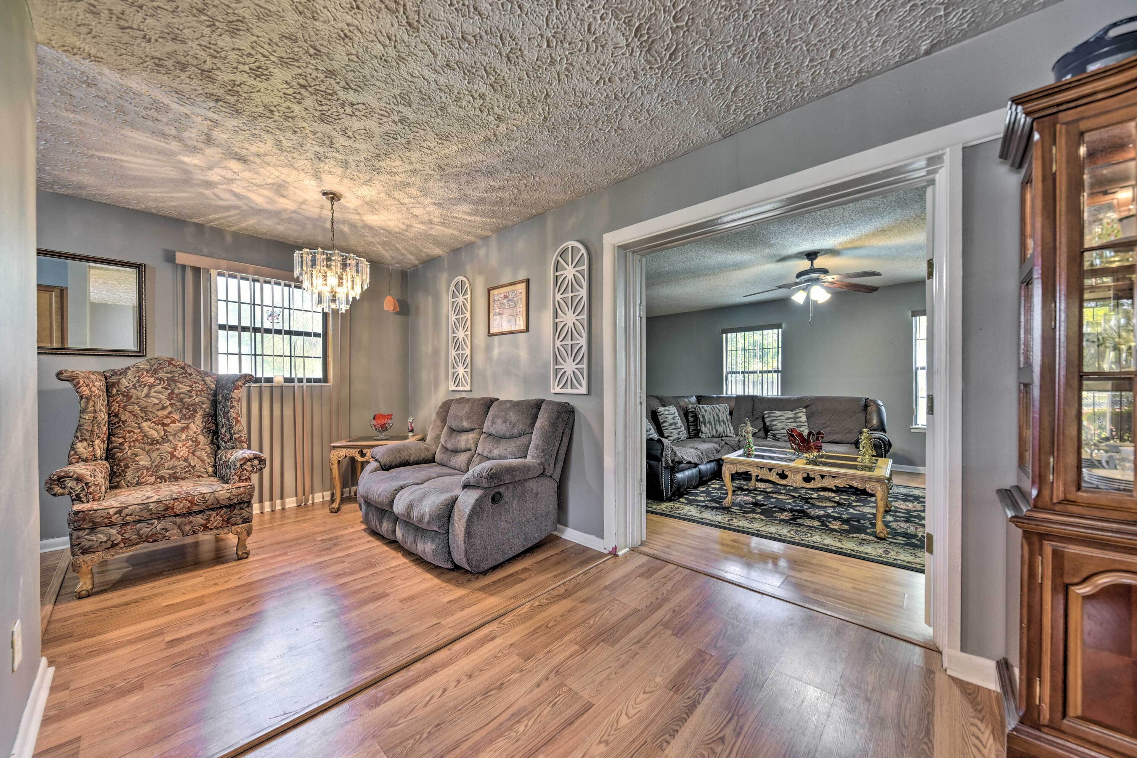 A spacious living room with hardwood flooring, a large gray sofa, a recliner, and a chandelier.