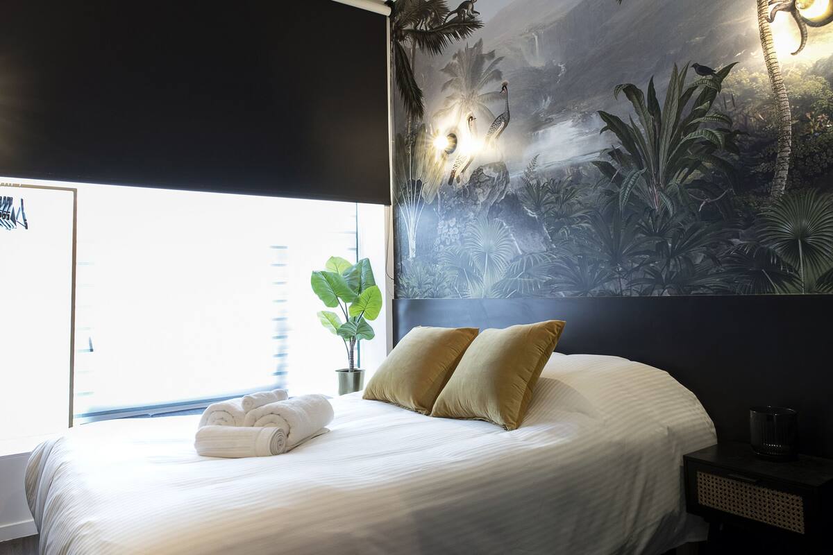 Ein ordentlich bezogenes Bett mit weißen Leinen und zwei goldenen Kissen, ein Blumentopf sowie ein Wandbild mit einer tropischen Szenerie.