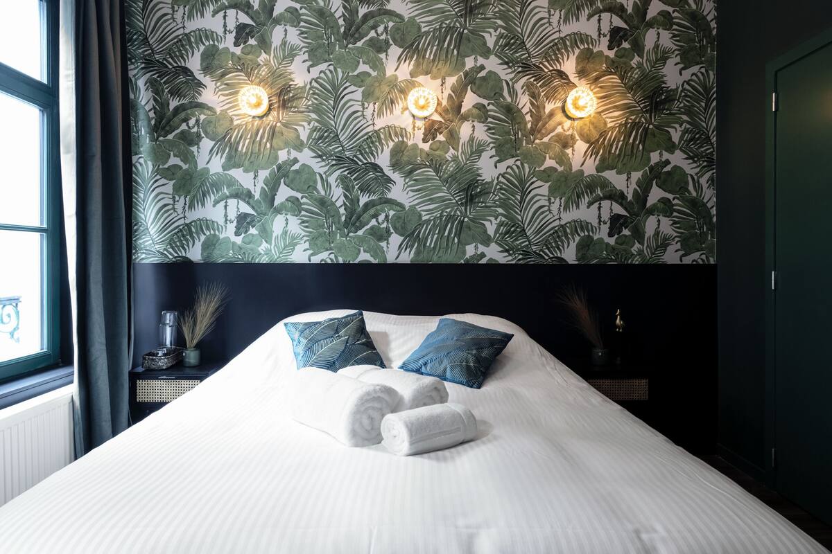 Ein ordentlich bezogenes Bett mit weißen Laken und zwei blauen Kissen, eine Wand mit einem Blattmuster und zwei Wandleuchten.