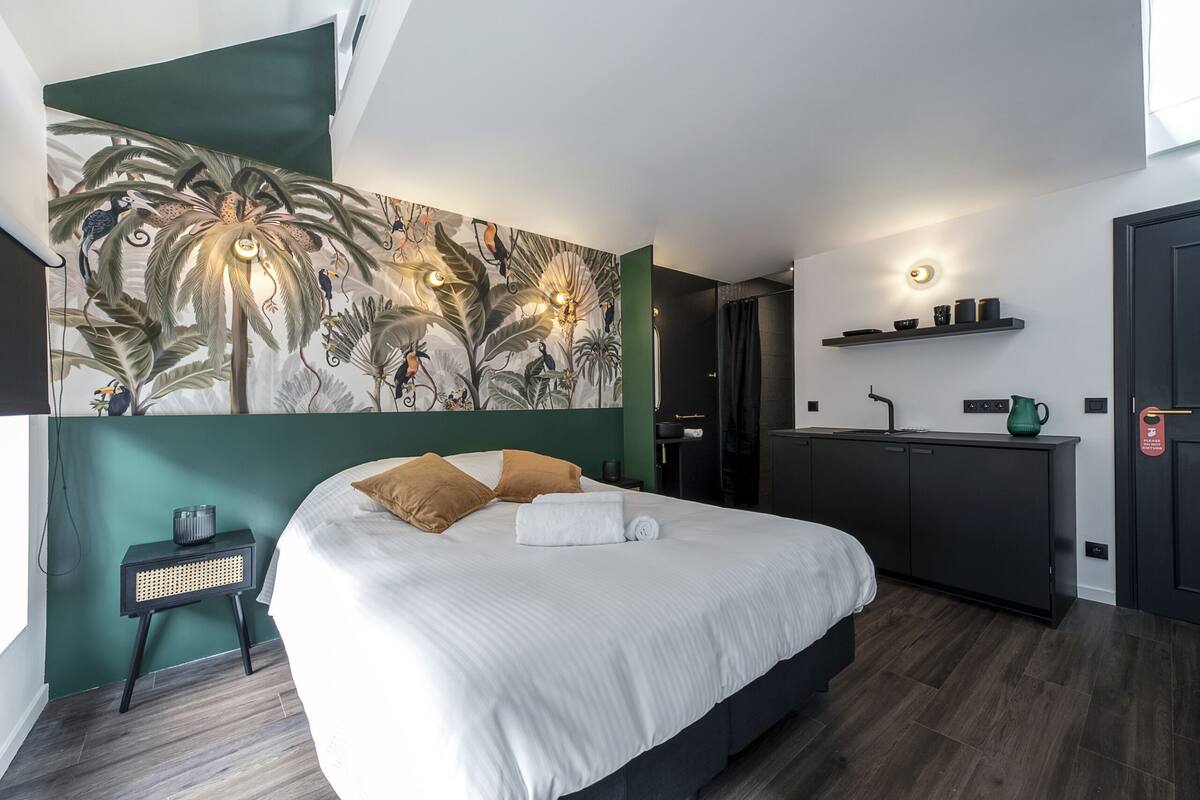 Ein modernes Hotelzimmer mit einem großen Bett, einem kleinen Nachttisch, einer Wandmalerei, einer Küchenzeile und einer Tür mit Griff.