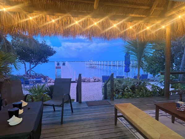 A wooden deck with outdoor seating, a thatched roof, and a view of the beach and pier.