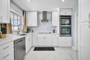A modern kitchen with white cabinets, a dishwasher, a double oven, and a window with curtains. - Stunning 4BR Hted Pool &Jacuzzi Close 2 the Beach (Deerfield Beach)