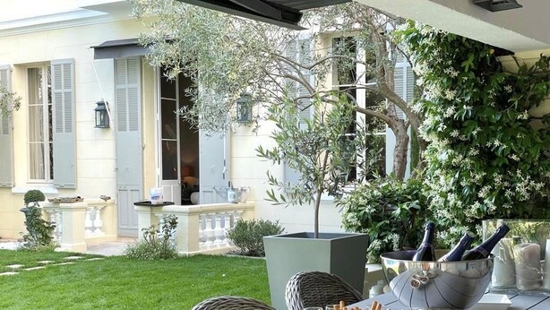 An outdoor dining area with a table set for a meal, surrounded by wicker chairs, a potted tree, and a view of a building with shutters.