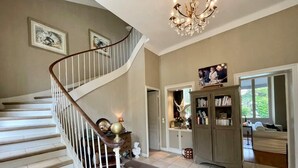 A spacious living area with a staircase, a chandelier, a bookshelf, and a window with a view.
