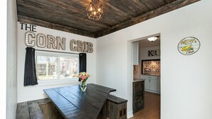 Una cocina rústica con una mesa de madera y bancos, una ventana con cortinas, y un letrero que dice "THE CORN CRIB".