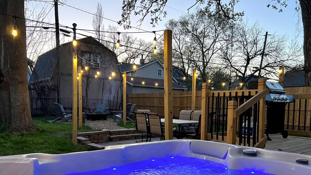 Un patio trasero con jacuzzi, terraza de madera, mobiliario exterior e iluminación con luces colgantes.