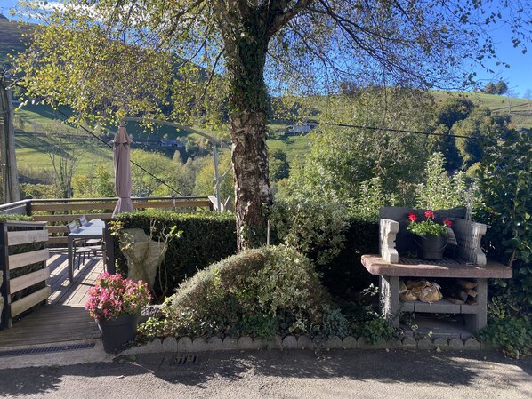 Une terrasse en bois avec un banc et une table, entourée de verdure et d’un arbre.