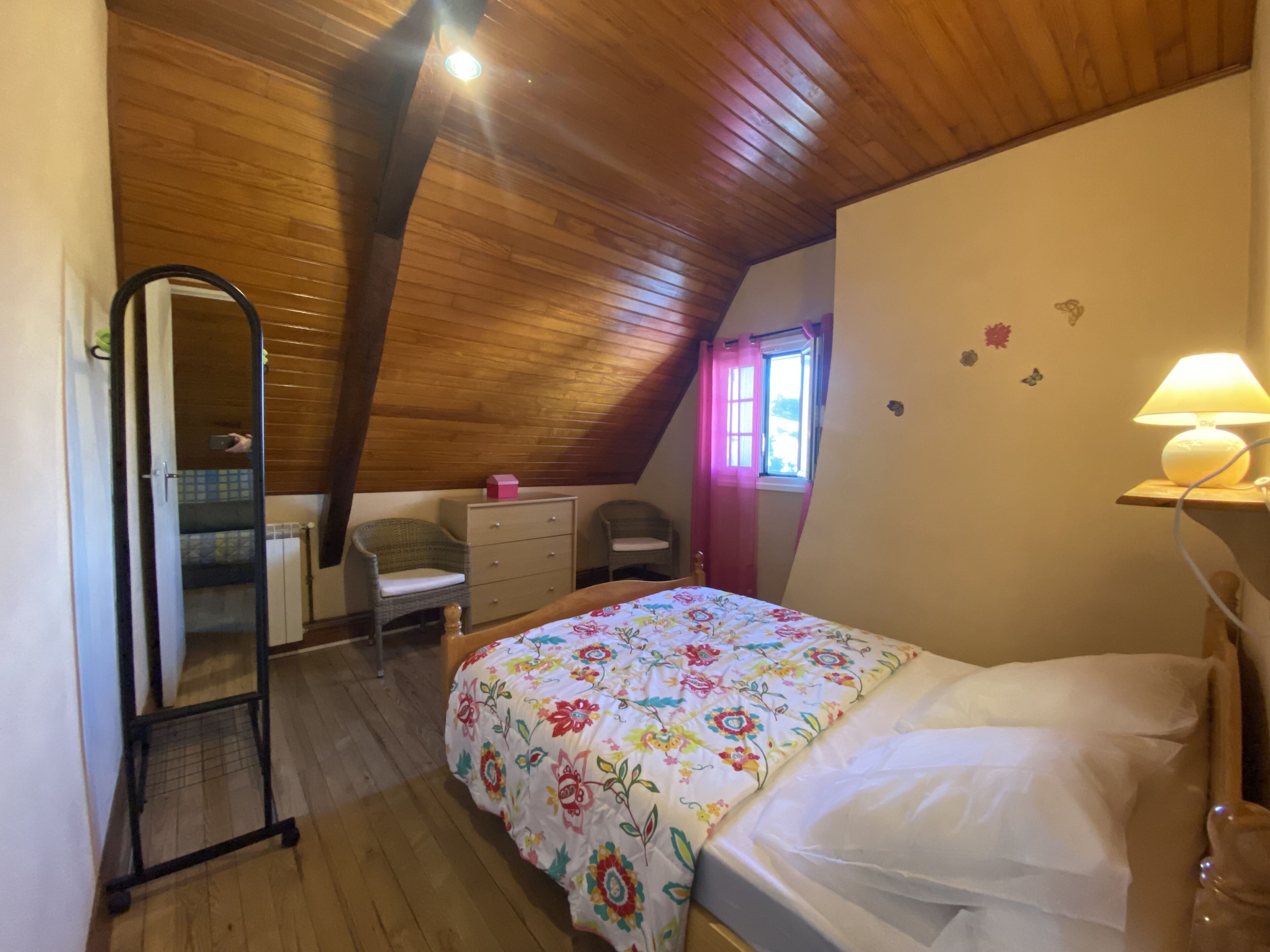 Une chambre avec un plafond en bois, un lit recouvert d’une couvre-lit à motifs floraux, un bureau avec une chaise, une commode et un miroir.
