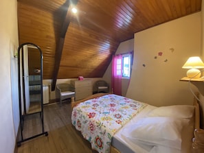 Quarto com teto de madeira, cama com colcha floral, escrivaninha com cadeira, cômoda e espelho.