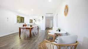 Ein geräumiger, helles Zimmer mit einem Holzespeisetisch und Stühlen, einem kleinen runden Tisch mit weißem Tuch und einem Korbstuhl mit Kissen.