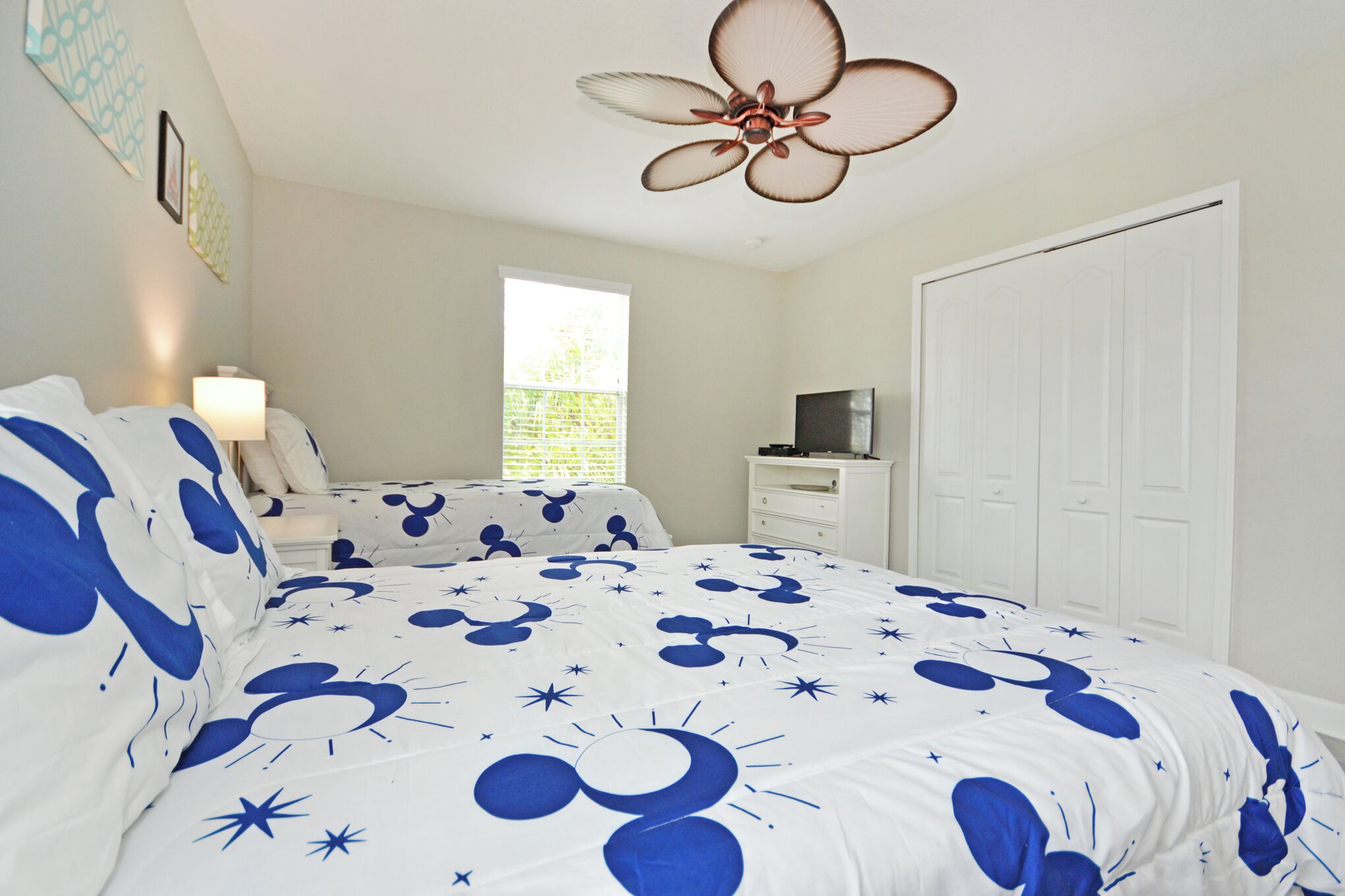 A bedroom with a bed featuring a white and blue Mickey Mouse themed bedspread, a ceiling fan, a window with blinds, a white dresser, and a white wardrobe.