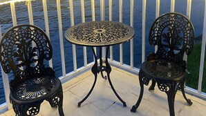 Un balcón con dos sillas de metal ornamentadas y una mesita, con vista a una masa de agua donde se ven barcos y un edificio al fondo.