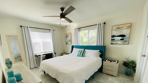 Un dormitorio con cama, escritorio, silla, mesita de noche, ventilador de techo y ventana con cortinas.