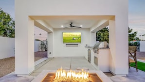 A modern outdoor kitchen area with a built-in grill, a flat-screen TV mounted on the wall, and a fire feature in the foreground. - Querencia  | Pool, Hot Tub, Putting Green, Fire Pit, TPC Golf | Scottsdale (Scottsdale)