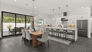 A modern kitchen with a central island, dark flooring, and a dining area with a glass door leading to an outdoor patio. - Querencia  | Pool, Hot Tub, Putting Green, Fire Pit, TPC Golf | Scottsdale (Scottsdale)