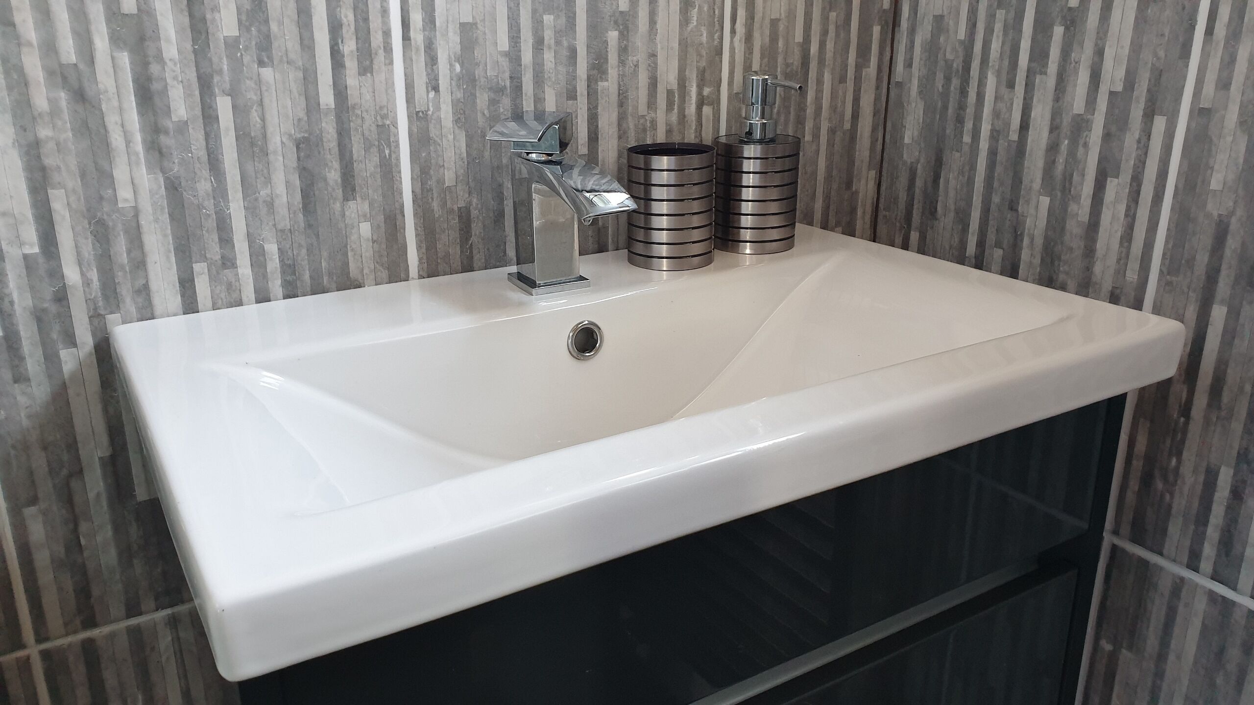 Un baño moderno con un lavamanos blanco, dos dispensadores metálicos de jabón y una pared con textura.
