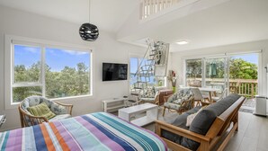 Une pièce spacieuse comprenant un lit, un bureau et une chaise. Un téléviseur est fixé au mur, et un balcon offre une vue sur les arbres.