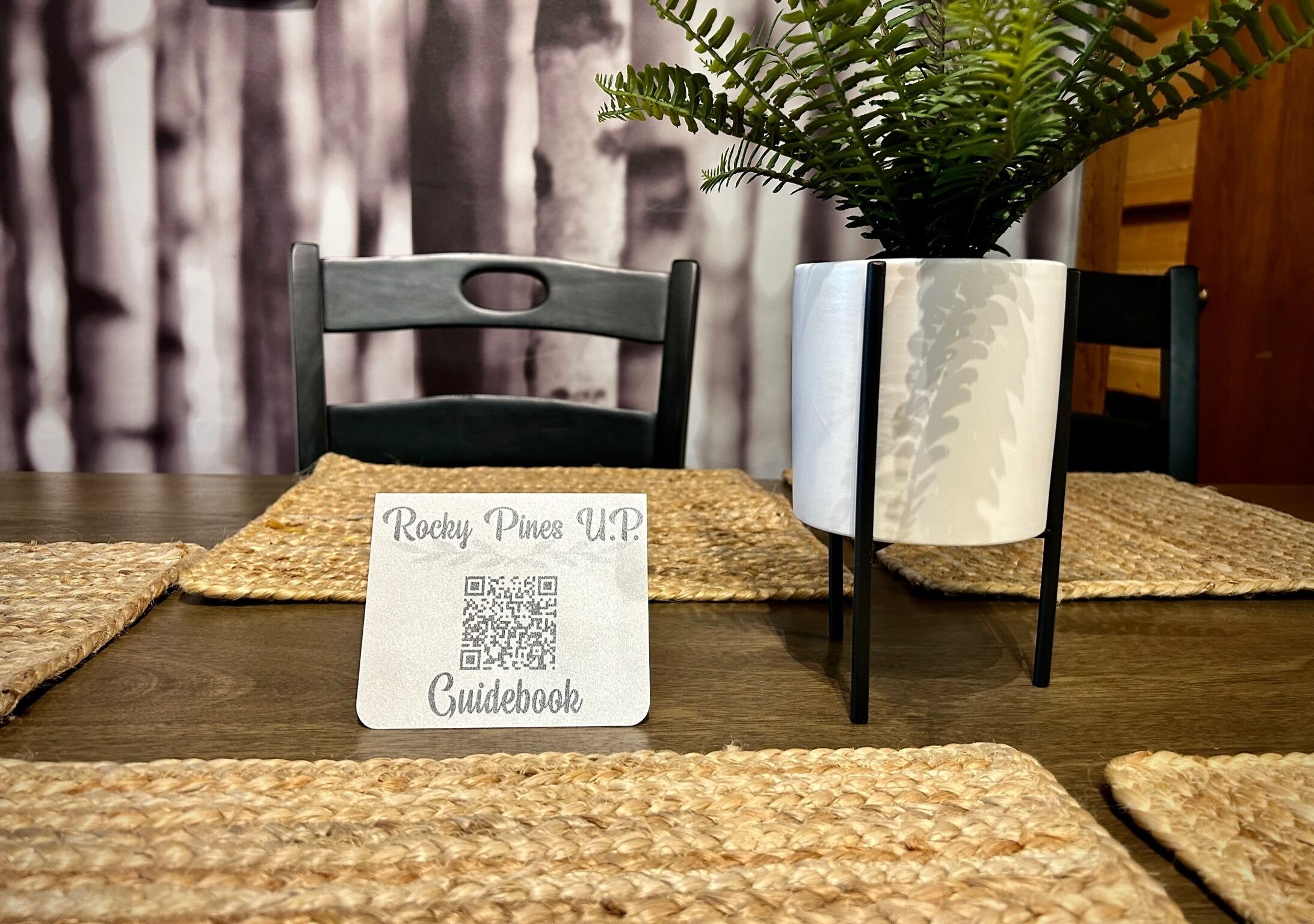 Een tafel met een placemat, een QR-code met de tekst 'Rocky Pines U.P. Guidebook' en een bloempot met een plantje.