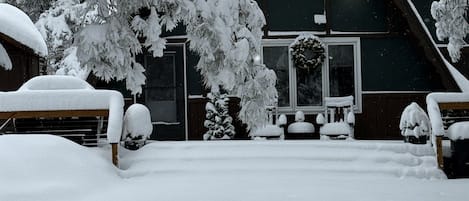 A snow-covered cabin with a steep roof, large windows, and a porch area.