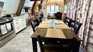 Een gezellig eetgedeelte met een houten tafel voor vier personen, omringd door stoelen. Links is een keuken zichtbaar met witte kasten en een magnetron.