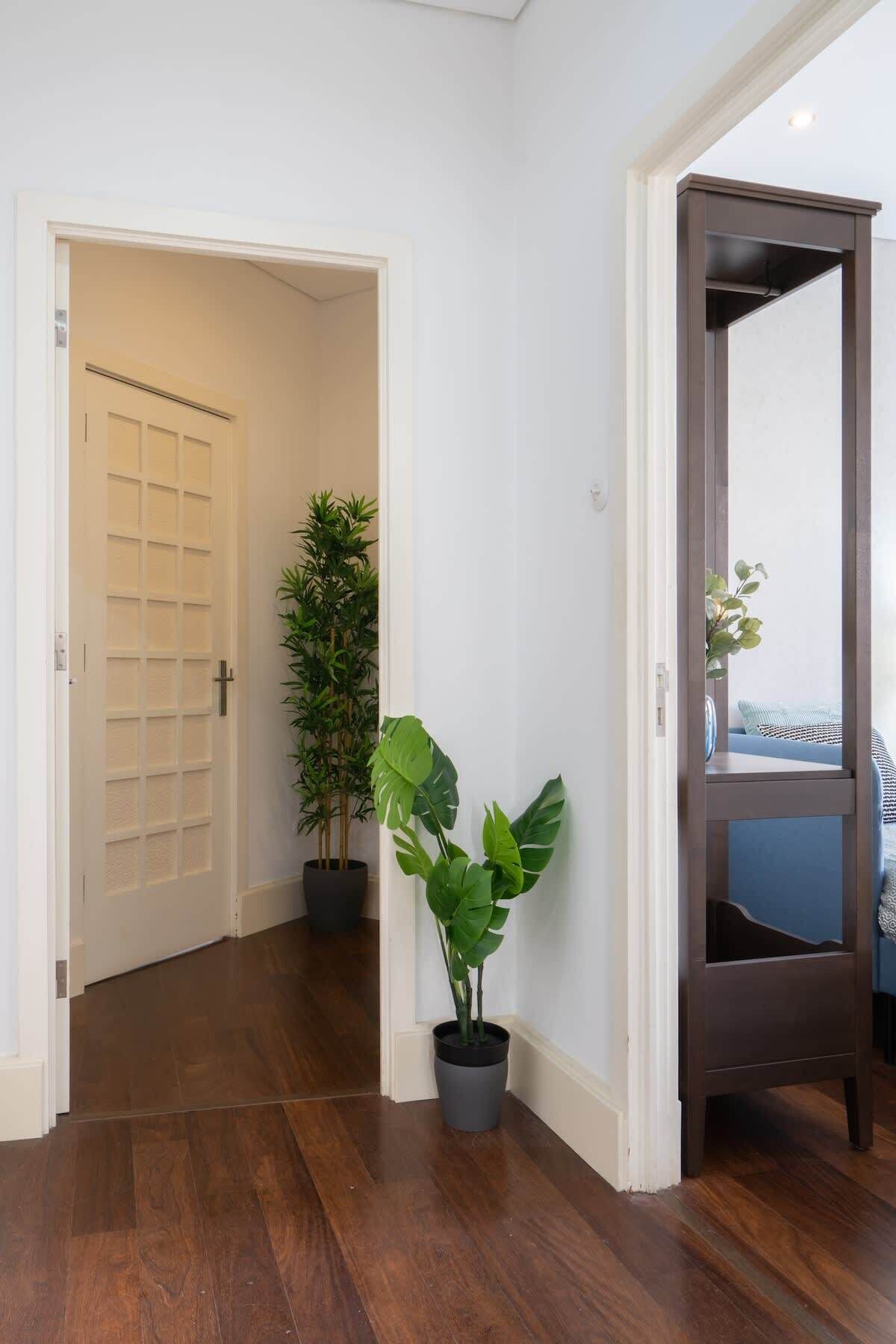 A hallway with wooden flooring, a white door, and a dark wooden door frame. There are two potted plants, one with broad leaves and another with slender leaves.