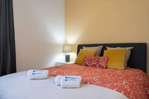 A neatly made bed with a red floral bedspread, two pillows, and two towels with the 'hostwise' logo. - Rustic Central Flat | Terrace by Host Wise (Porto)
