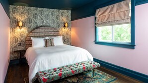Ein Schlafzimmer mit einem großen Bett, zwei Wandlampen, einem Fenster mit einer beigefarbenen Jalousie und einer gepolsterten Bank mit Blumenmuster.