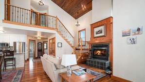 A spacious living area with a fireplace, wooden staircase, and a dining area with a kitchen in the background.