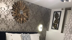 Ein ordentlich bezogenes Bett mit weißer Bettwäsche und einer schwarzen Tagesdecke, ein dekorativer Spiegel an der Wand und Tapete mit Muster.