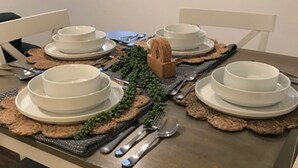 Ein Esstisch, gedeckt mit weißen Tellern, Schüsseln und Besteck, sowie einem kleinen Holzklotz und grünen Dekorationselementen.