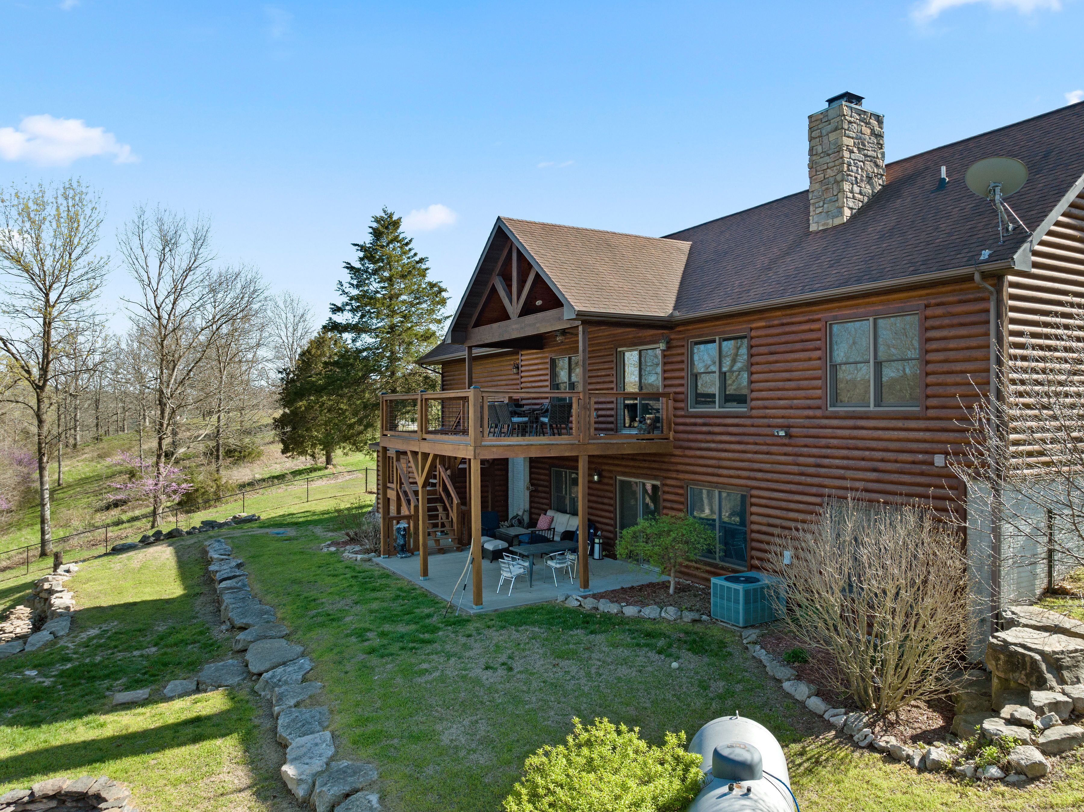 A log cabin with a two-story deck, outdoor seating, and a stone retaining wall.