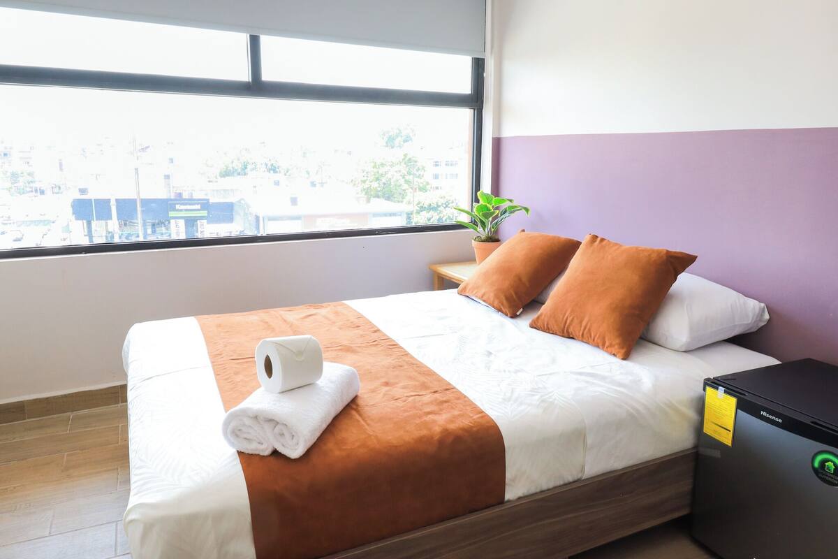 Ein ordentlich gemachtes Bett mit einer braun-weißen Tagesdecke, zwei orangefarbenen Kissen und einem aufgerollten weißen Handtuch. Auf einem Nachttisch neben dem Bett steht eine kleine Topfpflanze.