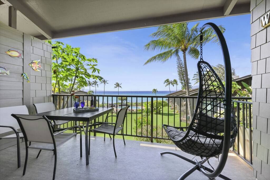 A balcony with a table and chairs, a hanging chair, and a view of the ocean and palm trees.