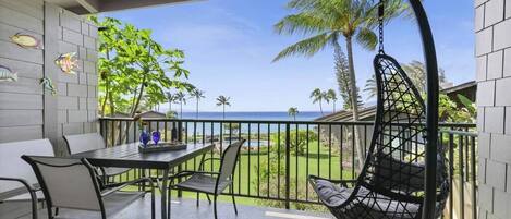 A balcony with a table and chairs, a hanging chair, and a view of the ocean and palm trees.