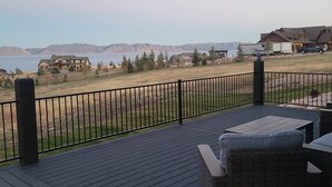 Una terraza con vistas a un lago y montañas, con mobiliario exterior y barandilla metálica.