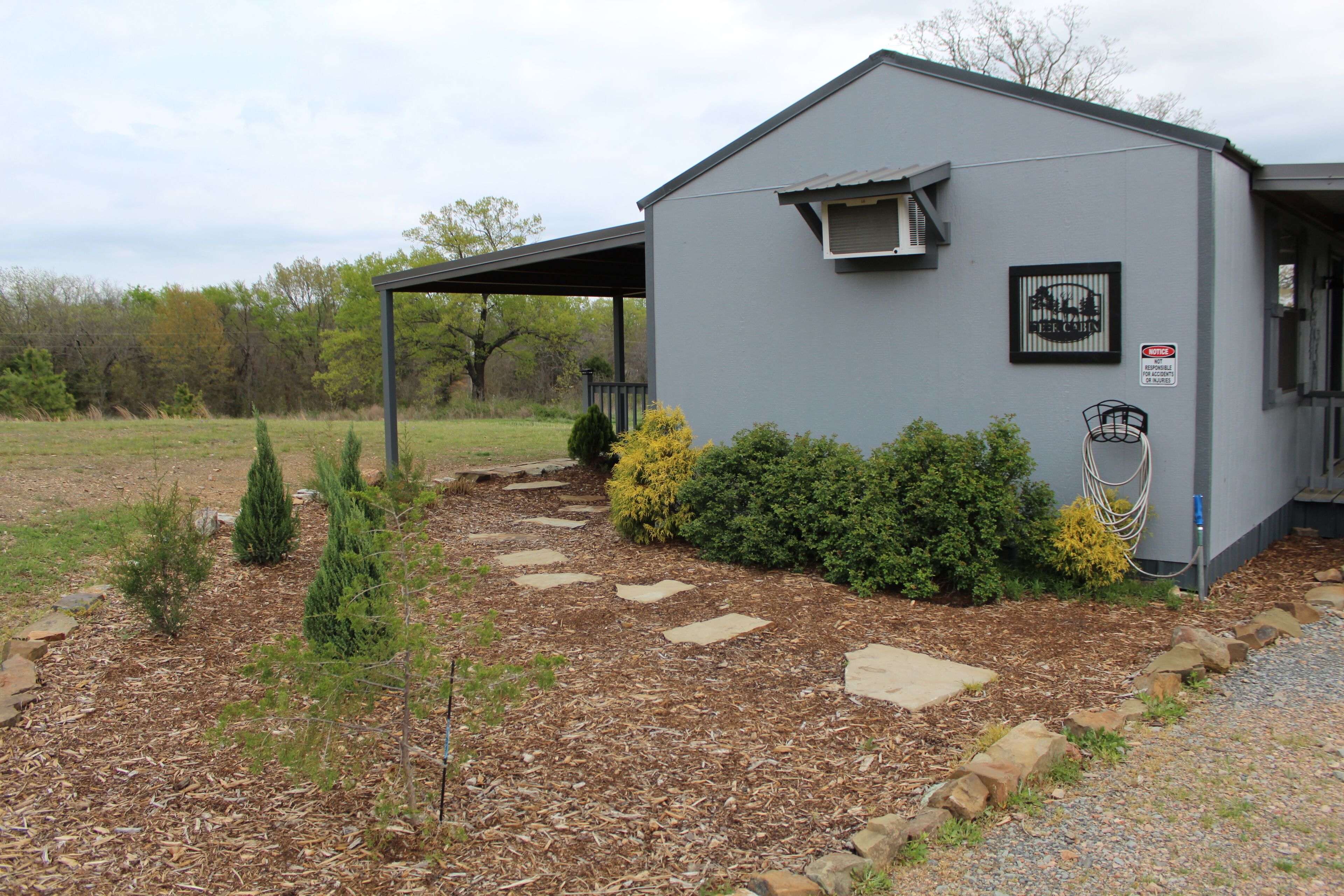 A small, single-story building with a grey exterior, a covered porch, and a landscaped garden with various shrubs and a stone pathway.