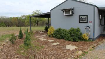 A small, single-story building with a grey exterior, a covered porch, and a landscaped garden with various shrubs and a stone pathway.