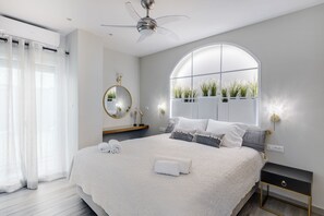 Un dormitorio moderno con una cama grande, un ventilador de techo, un espejo redondo y una ventana con plantas.