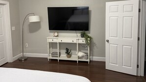 Ein Schlafzimmer mit einem Bett, einem an einer weißen Kommode angebrachten Fernseher, einer Stehlampe und einer Tür.