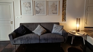 Un salon avec un canapé gris, deux coussins, une petite table ronde sur laquelle se trouve une lampe, et trois reproductions botaniques encadrées accrochées au mur.