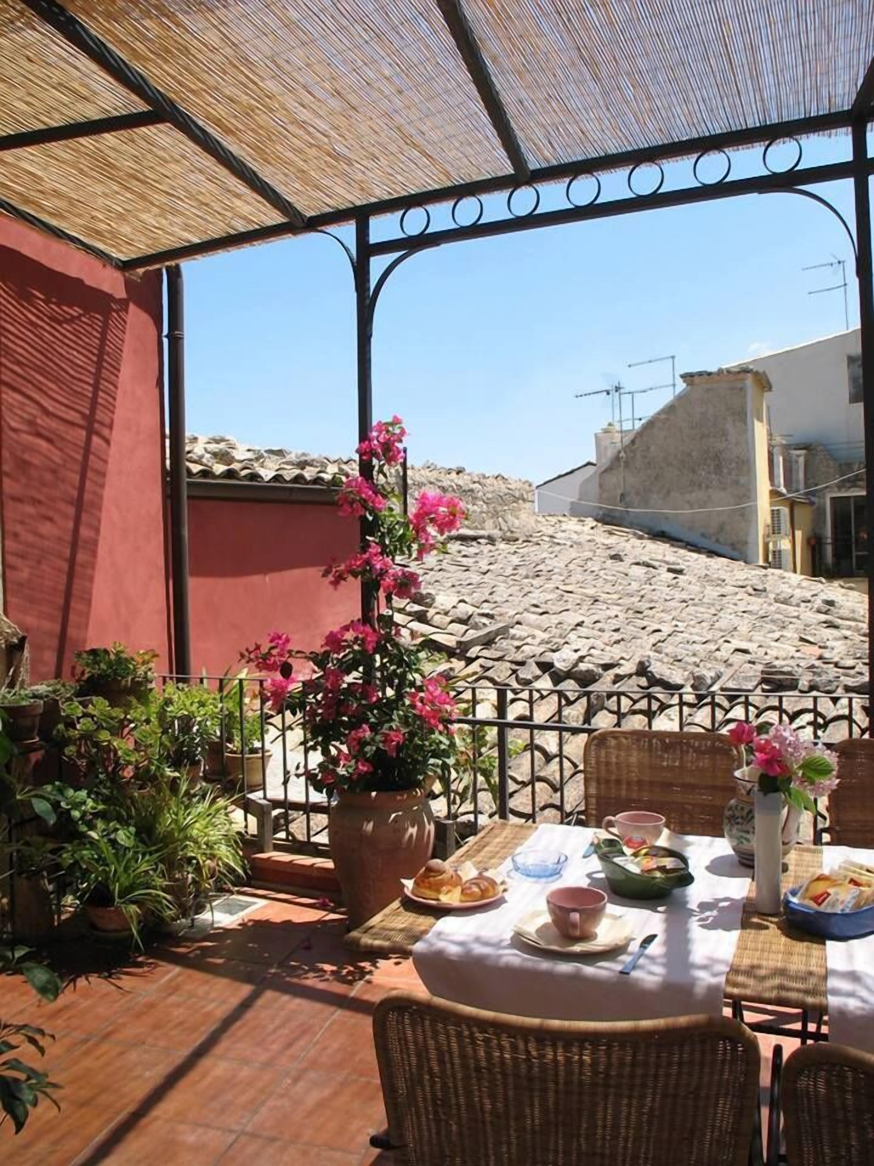 An outdoor dining area with a table set for two, surrounded by wicker chairs, potted plants, and a pergola overhead.